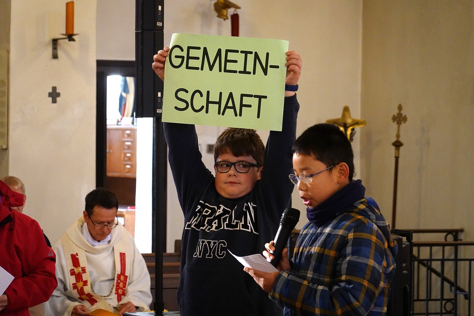 Ein Junge ließt eine Fürbitte, ein anderer hält ein Schild mit "Gemeinschaft" über seinem Kopf